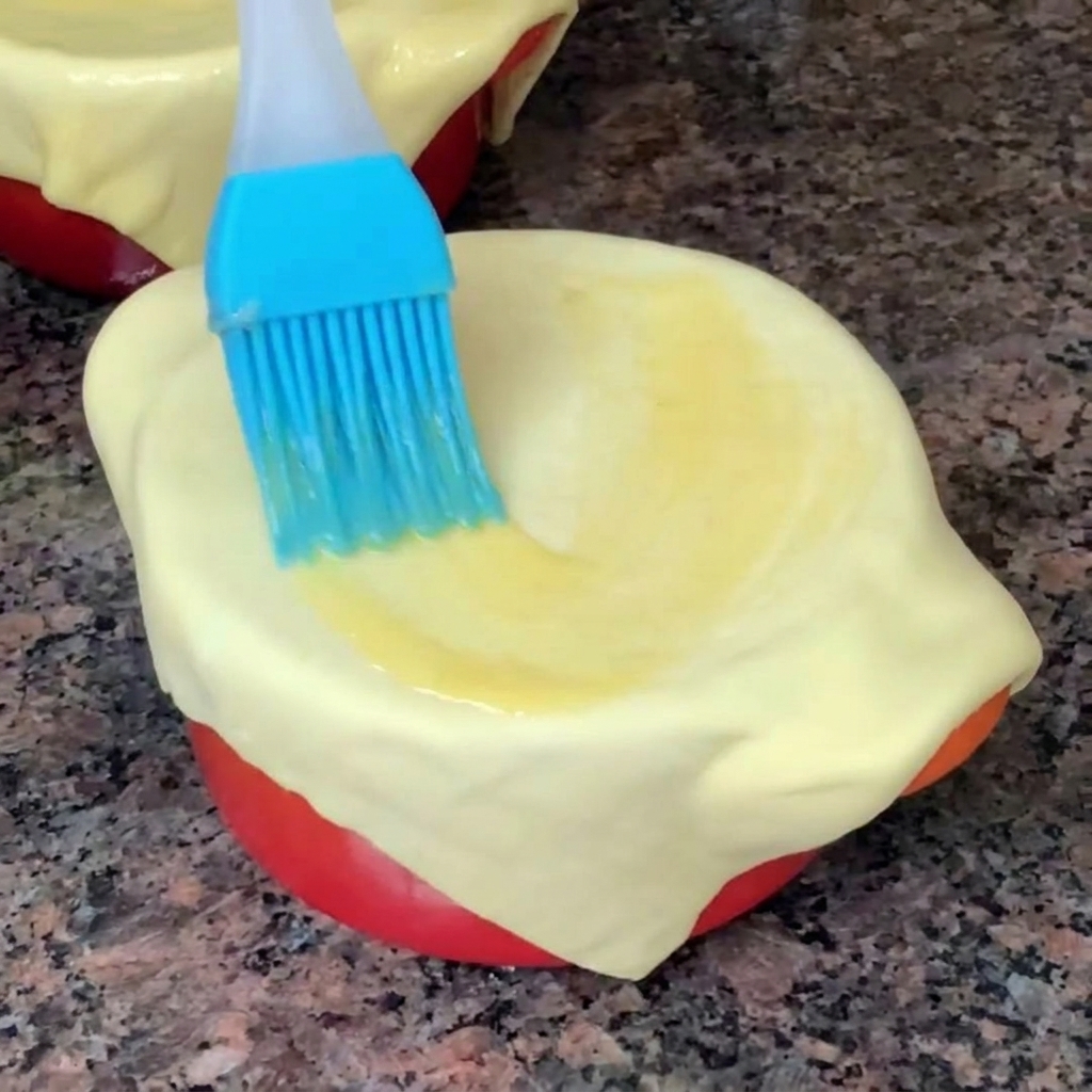 Primer plano de una brocha de silicona azul extendiendo huevo batido sobre una hoja de hojaldre cruda.
