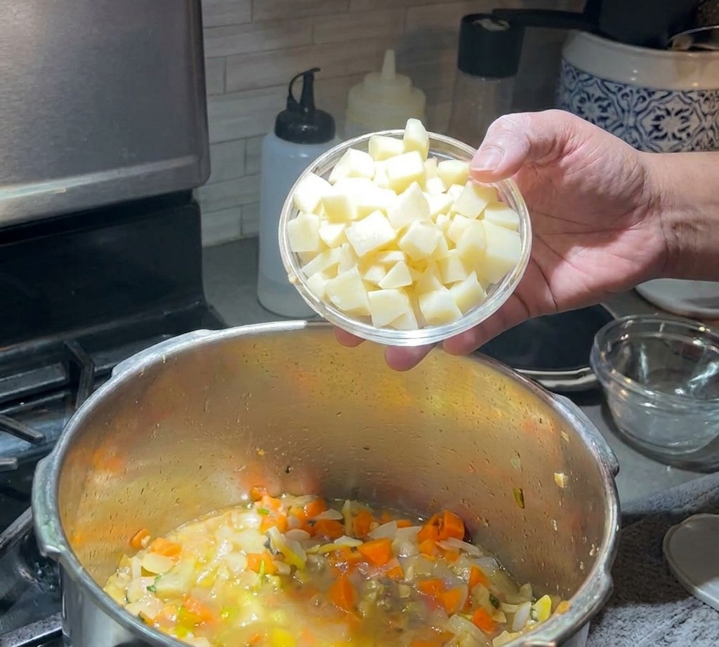 Una mano vertiendo un pequeño tazón de papas en cubos en una olla de sopa a fuego lento.