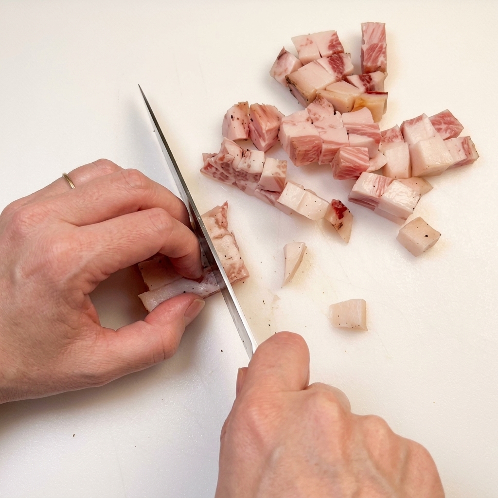 Mano con un cuchillo cortando bacon en dados uniformes sobre una tabla.