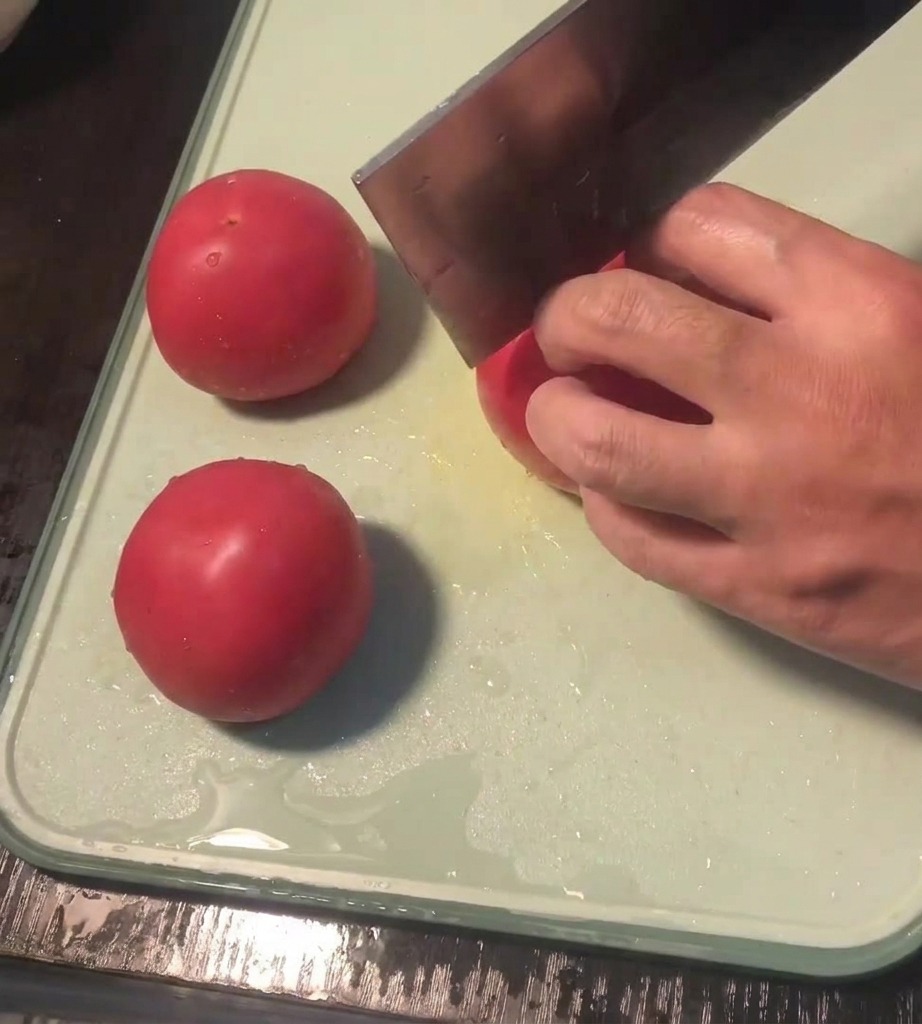 Primer plano de una mano usando un cuchillo para hacer una cruz en la piel de un tomate fresco sobre una tabla de cortar.