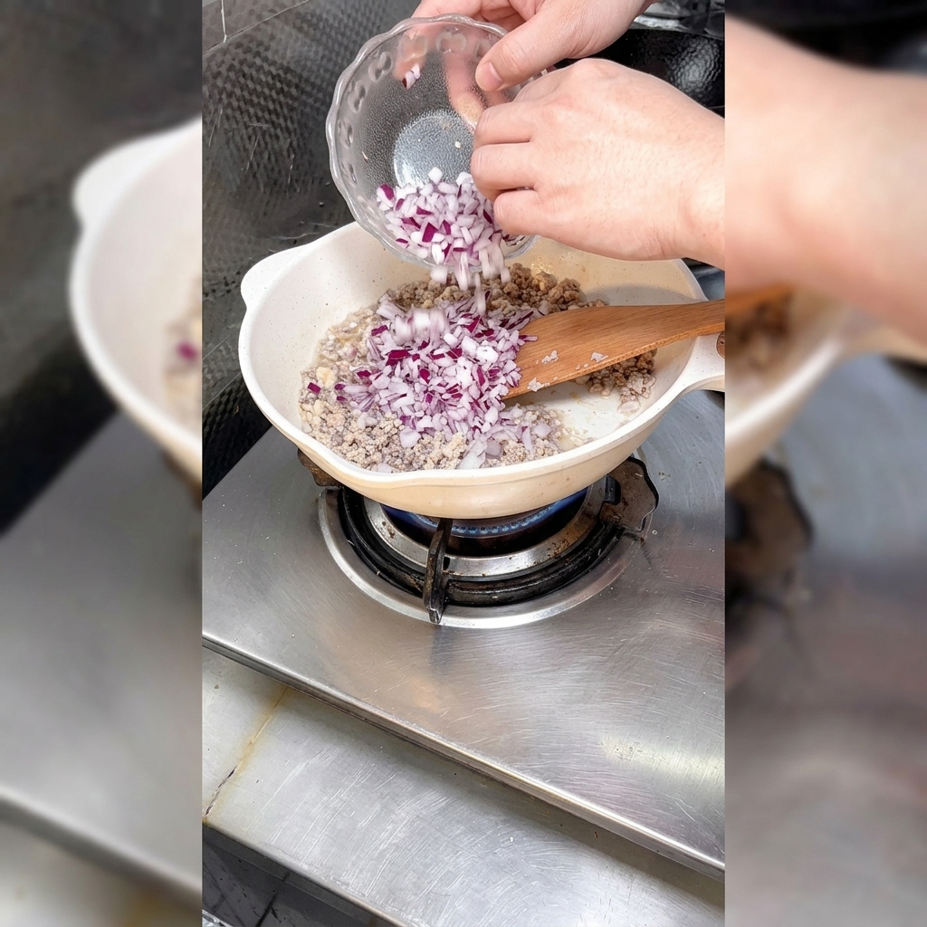 Cebollas picadas siendo añadidas a una sartén llena de carne picada dorada.