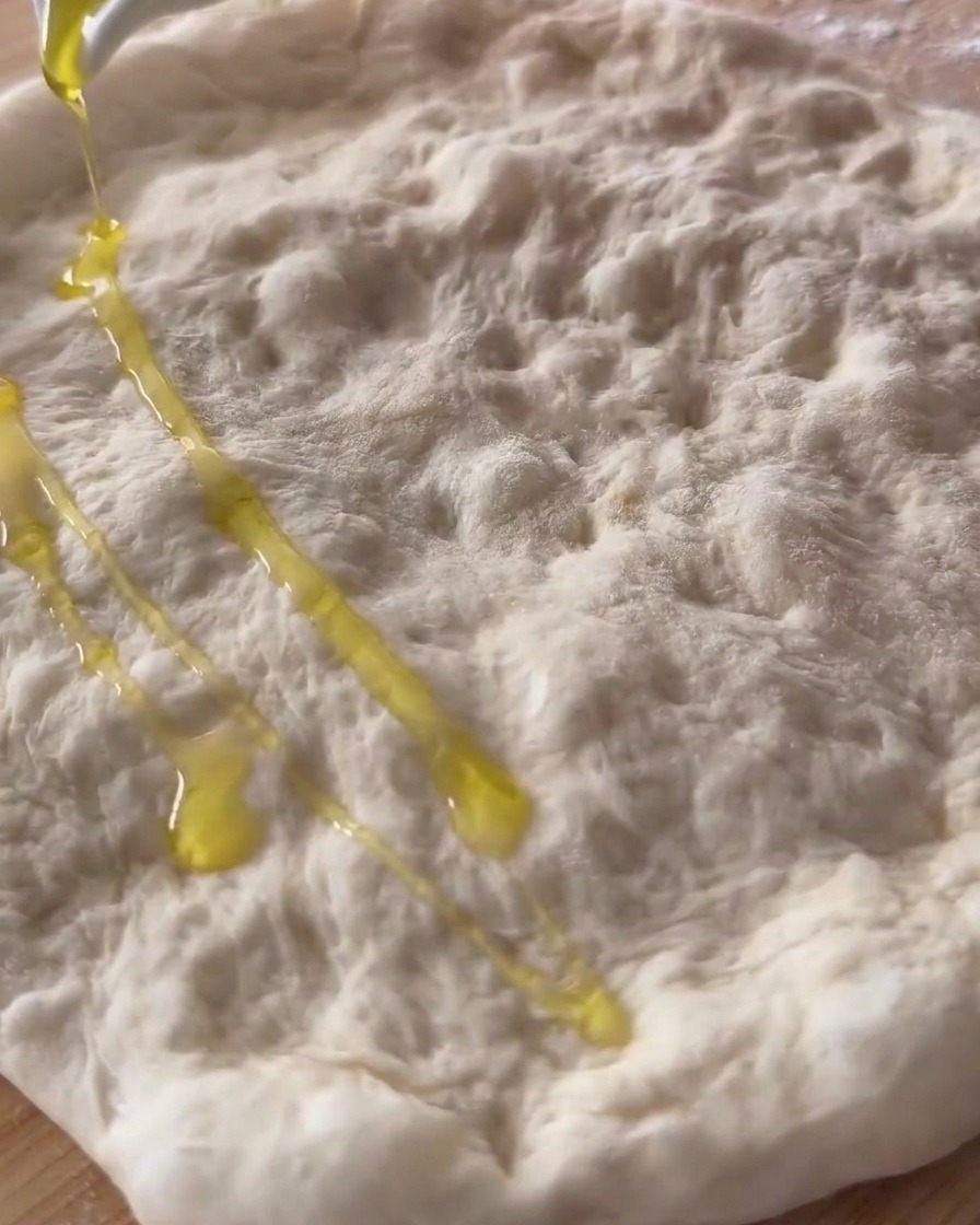 Aceite de oliva siendo vertido sobre una masa de pizza cruda y estirada.