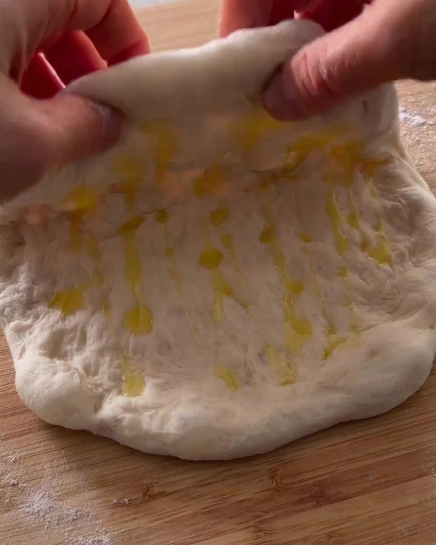 Manos doblando un trozo de masa de pizza con aceite por la mitad sobre una superficie de madera.