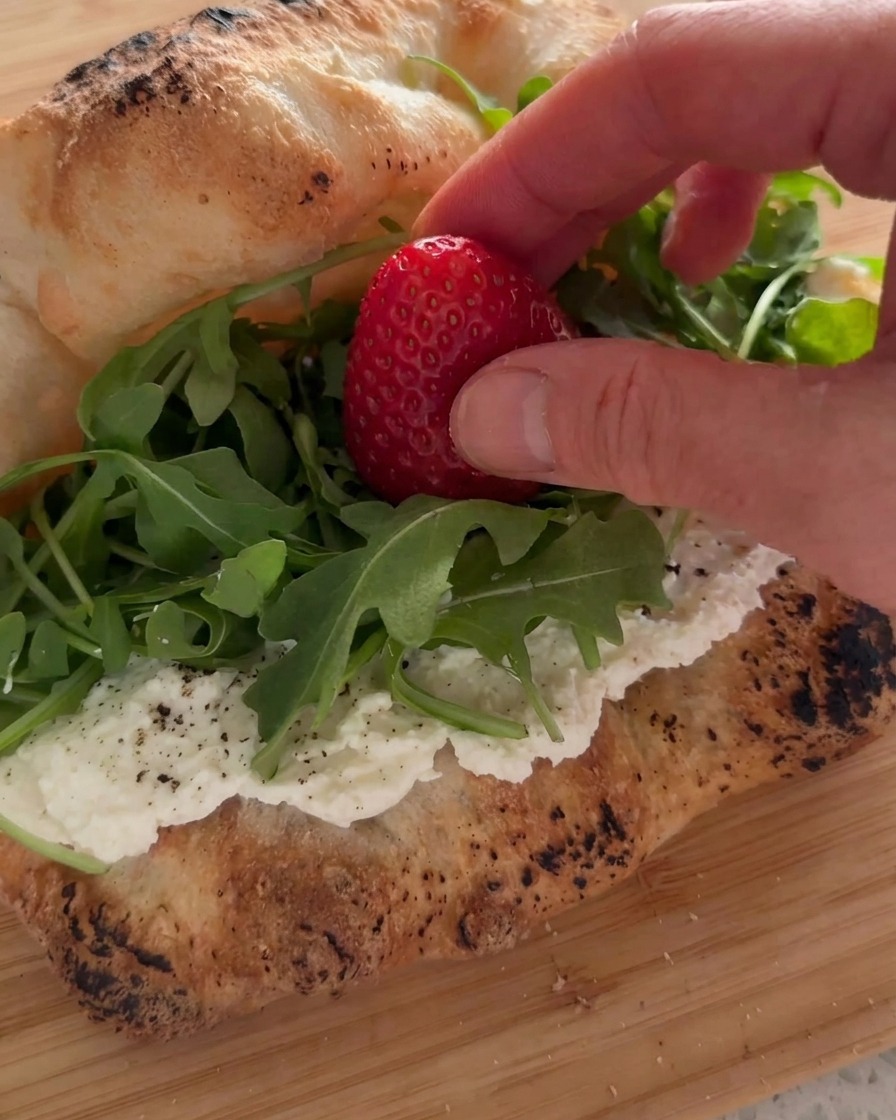 Una mano colocando una fresa fresca sobre una cama de rúcula verde dentro de un sándwich cubierto de ricotta.