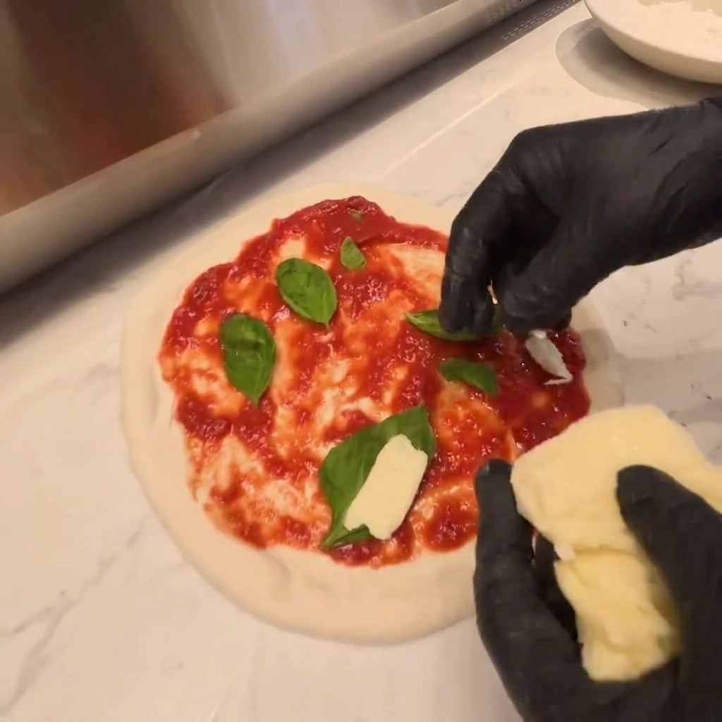 Trozos de queso mozzarella fresco siendo colocados sobre una capa de rúcula en masa de pizza cruda.