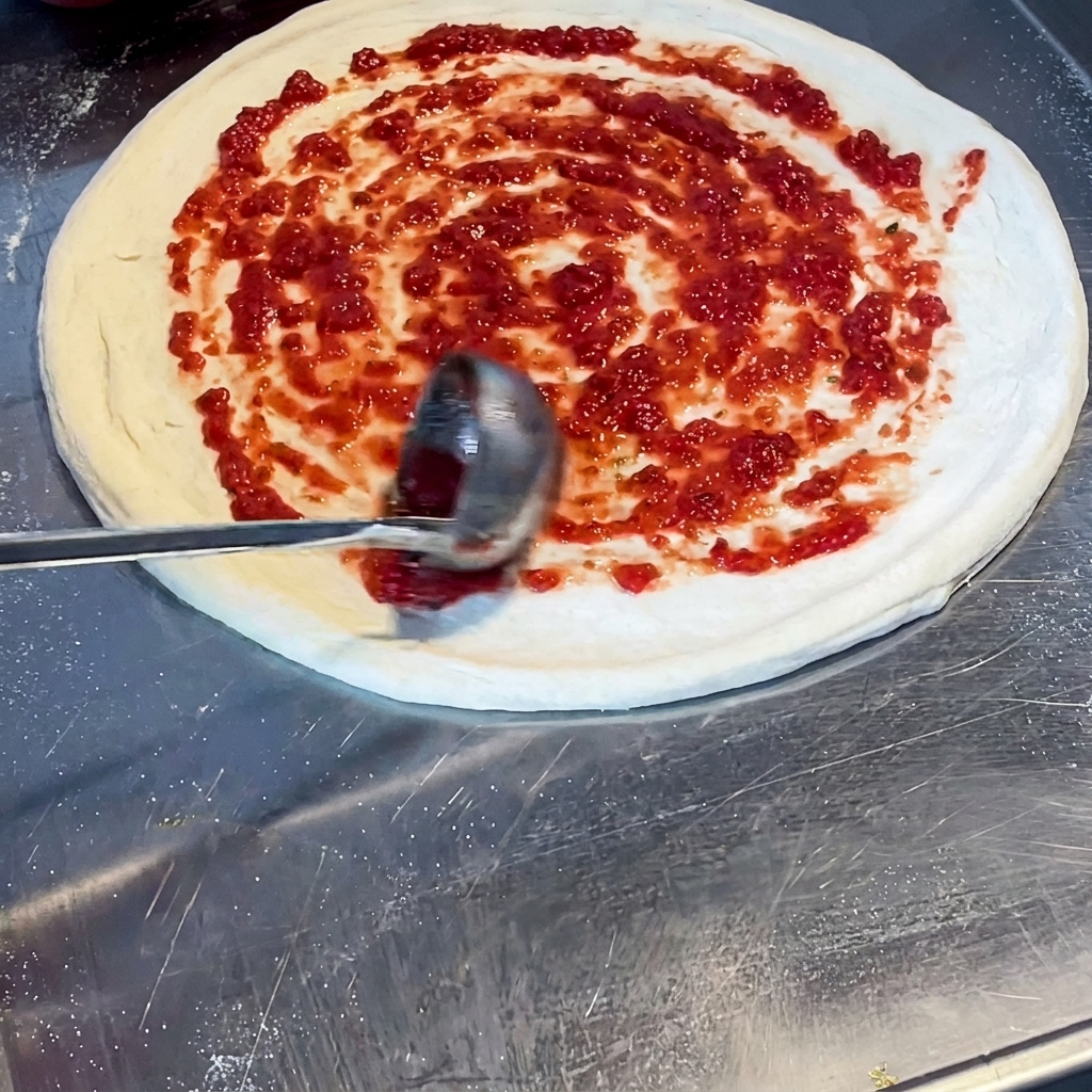 Un cucharón esparciendo salsa de tomate de un rojo vibrante en forma de espiral sobre la masa de pizza cruda.