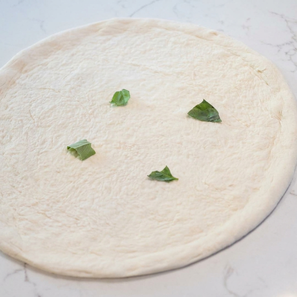 Una base de masa de pizza estirada cruda cubierta con hojas de albahaca verde dispersas.