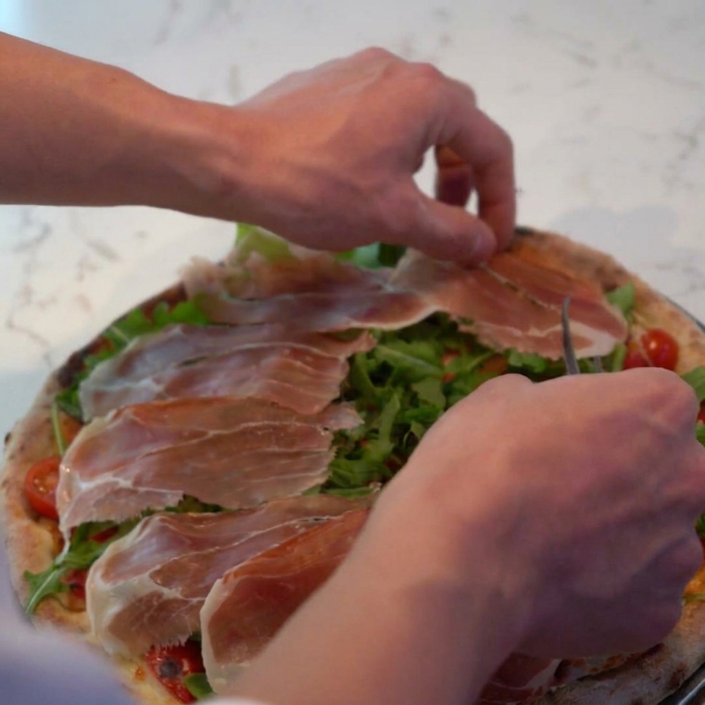 Queso parmesano rallado siendo espolvoreado sobre la pizza, cubriendo el prosciutto y la rúcula.
