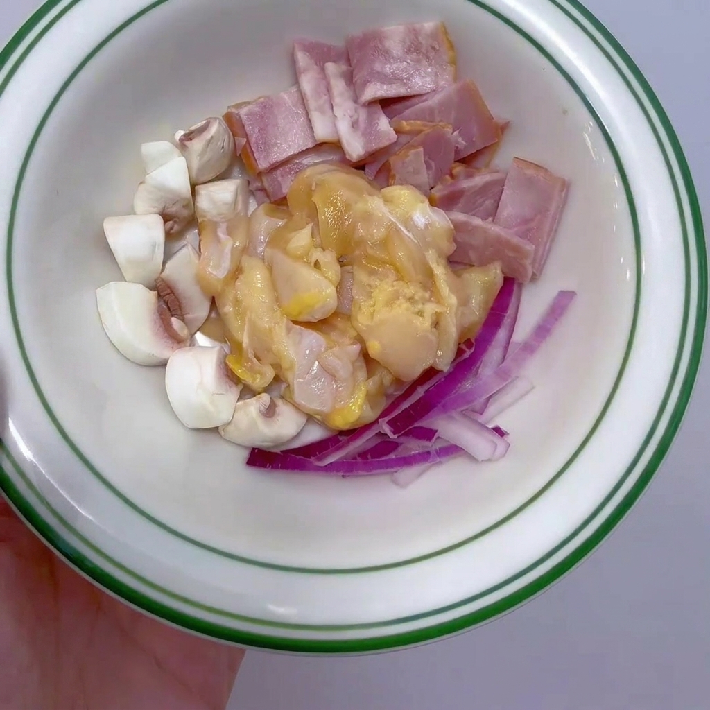 Un pequeño tazón con pechuga de pollo cortada en cubos, tiras de tocino, champiñones en cuartos y cebolla morada en tiras finas.