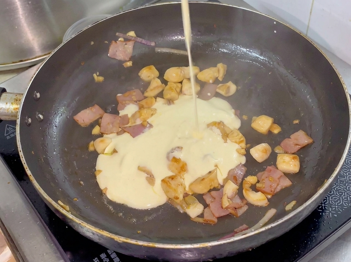Crema vertiéndose desde una jarra pequeña hacia la sartén con el pollo, tocino y vegetales.
