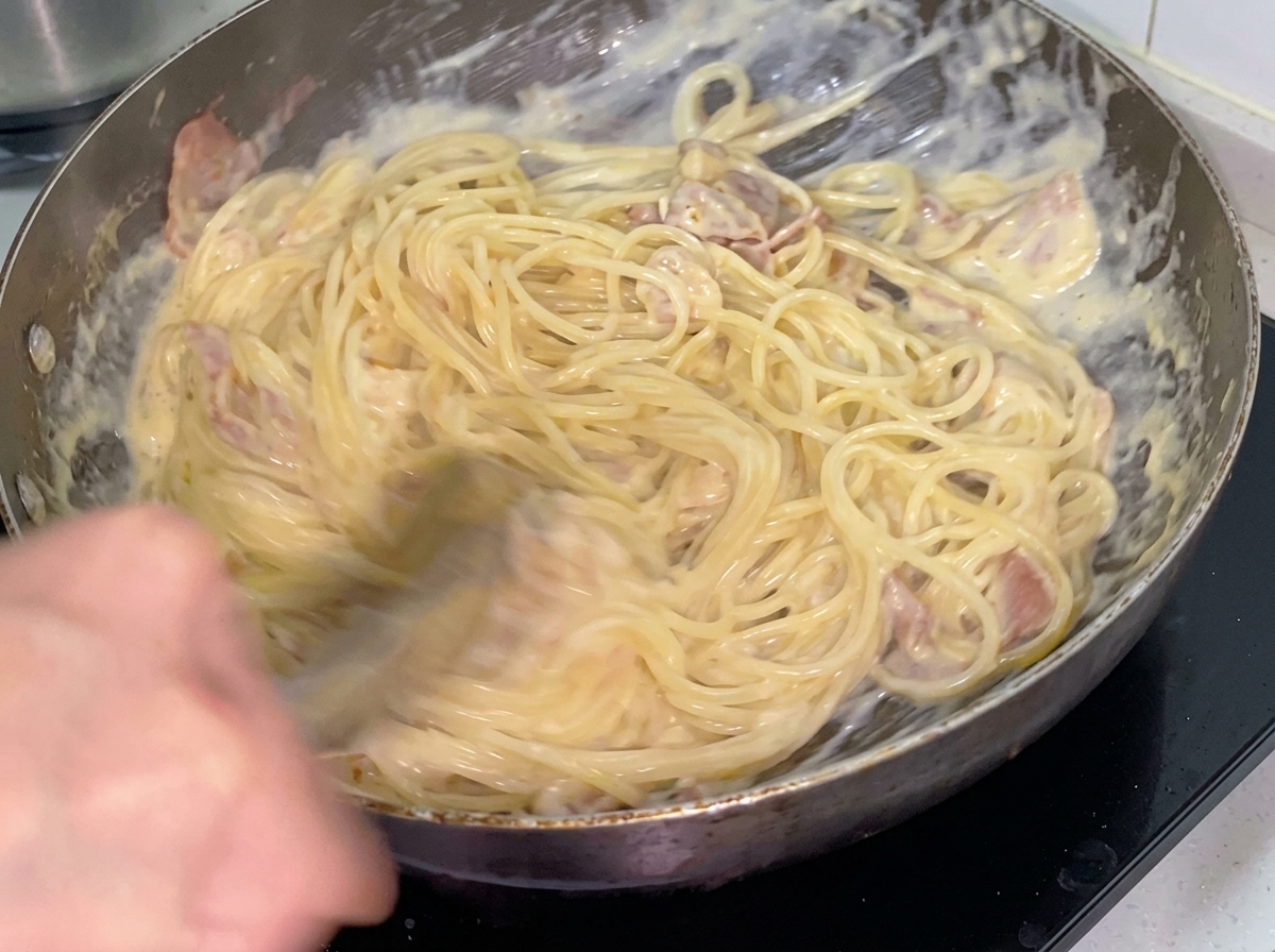 Espaguetis cremosos siendo mezclados con el pollo y tocino usando pinzas metálicas.