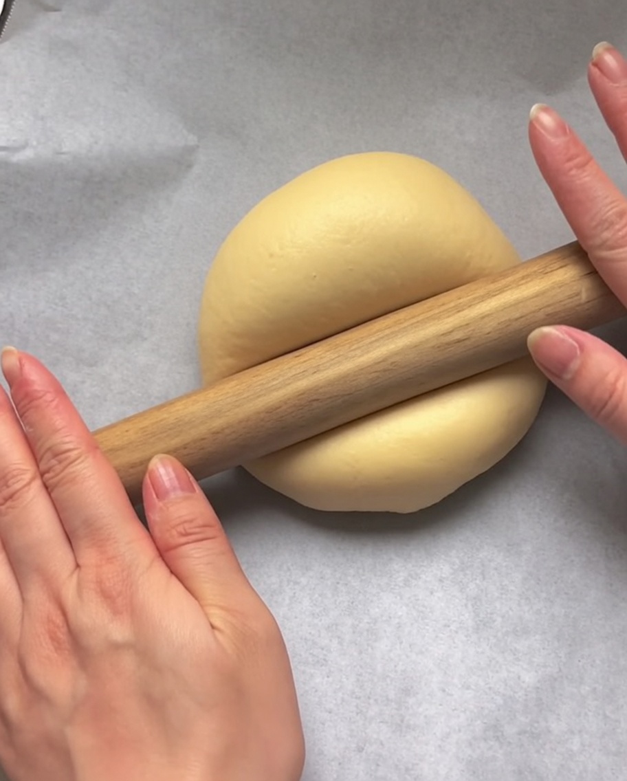 Manos usando un rodillo de madera para aplanar una bola de masa cruda en forma circular sobre papel pergamino blanco.