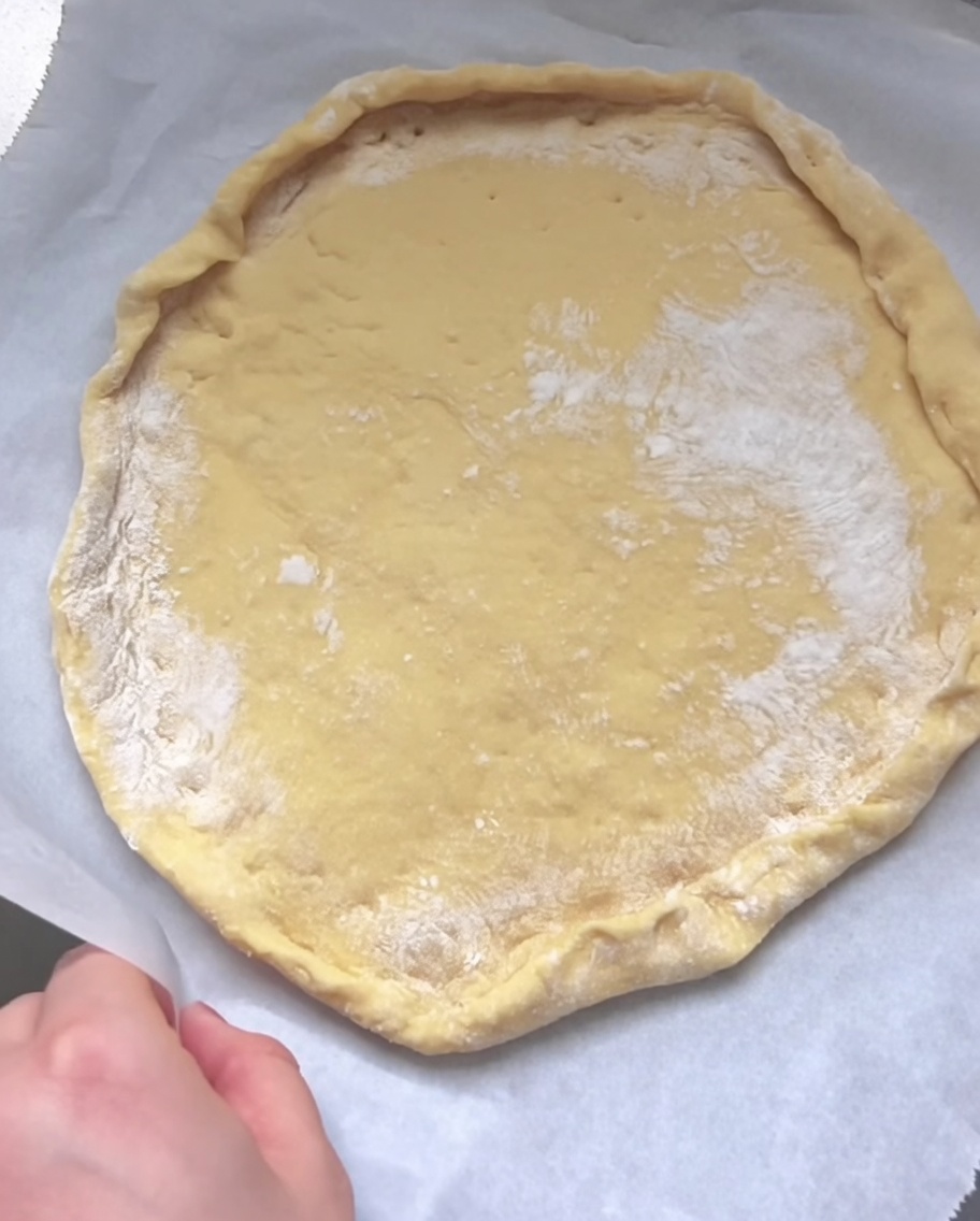 Una mano doblando y pellizcando el borde de la masa cruda aplanada para formar un borde elevado sobre papel pergamino.