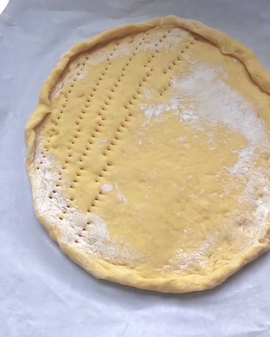Una masa circular cruda para pizza sobre papel pergamino con pequeños agujeros perforados uniformemente a lo largo de su superficie plana central.