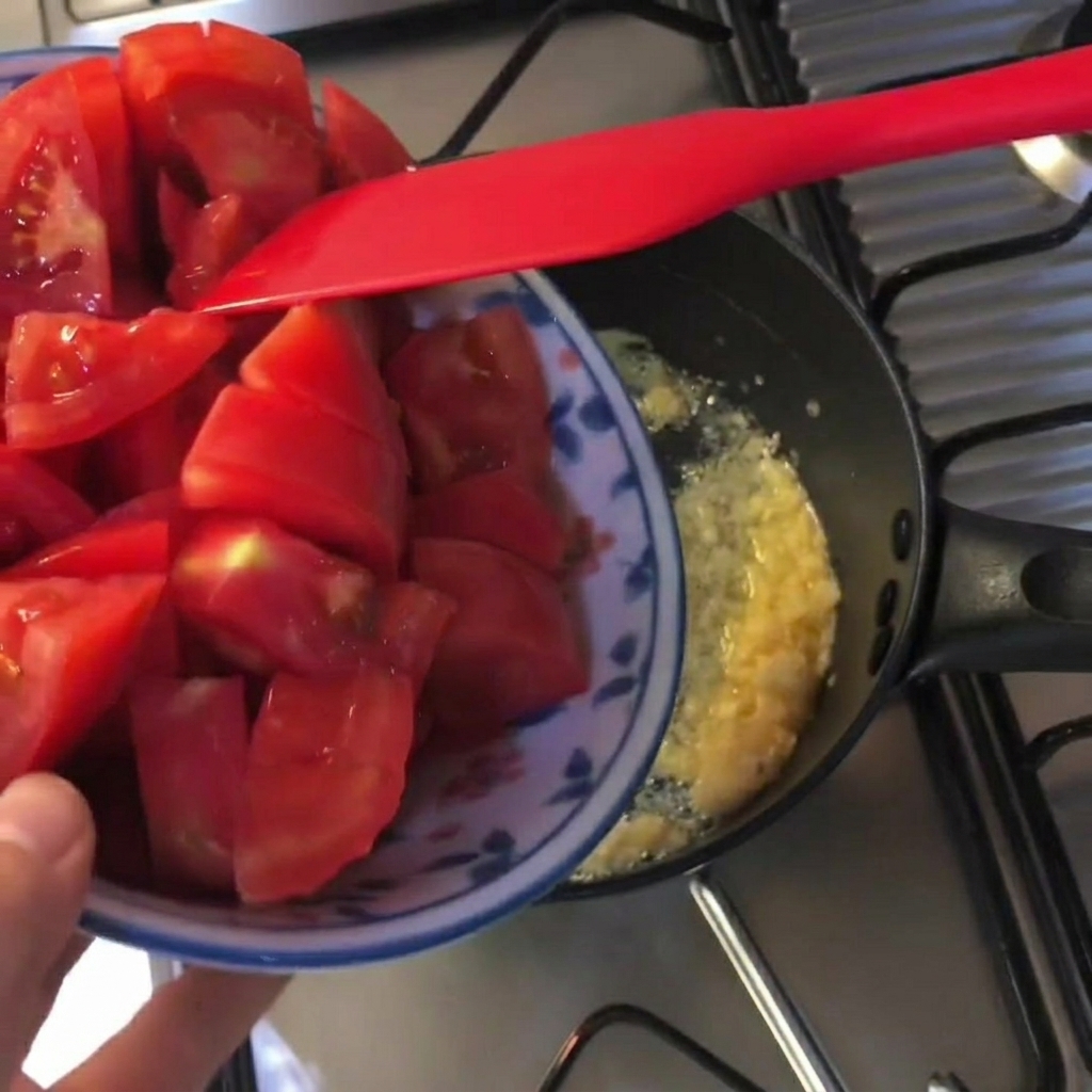 Trozos de tomate rojo fresco siendo agregados a una olla negra con mantequilla derretida y ajo picado.