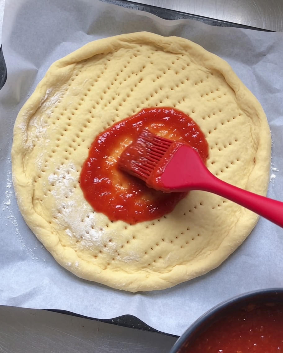 Salsa de tomate roja siendo esparcida uniformemente sobre una base redonda de masa de pizza cruda con agujeros.