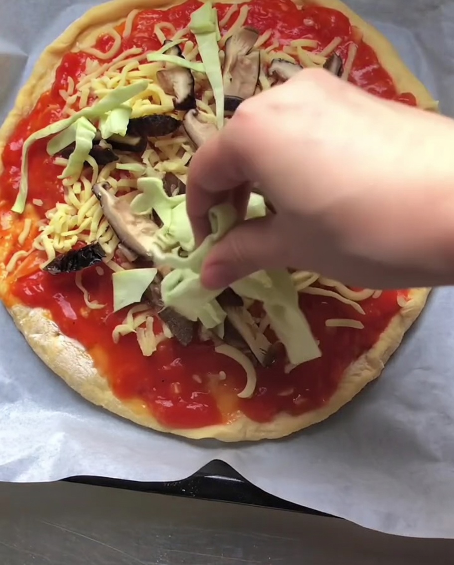 Una mano colocando repollo verde crudo rallado sobre una pizza con una capa de salsa de tomate, queso y champiñones en rodajas.
