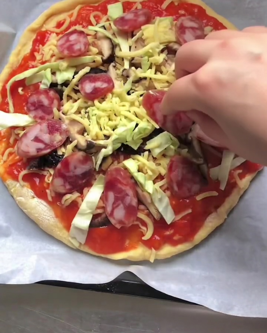 Una mano colocando rodajas de salchicha italiana roja en una pizza cruda cubierta con salsa, queso y verduras.