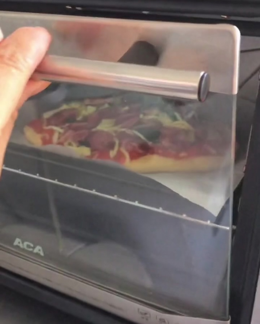 Abriendo la puerta de un pequeño horno de mostrador para colocar adentro una pizza cruda armada sobre una bandeja para hornear.