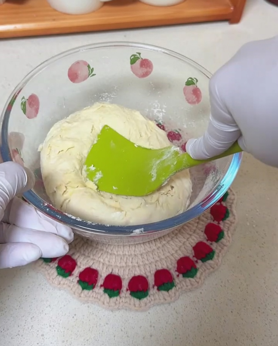 Manos enguantadas usando una espátula de silicona verde para mezclar una masa rústica dentro de un bol de vidrio.
