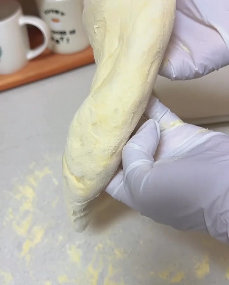 Manos enguantadas sosteniendo un trozo de masa suave verticalmente, permitiendo que se estire hasta formar una base de pizza fina.
