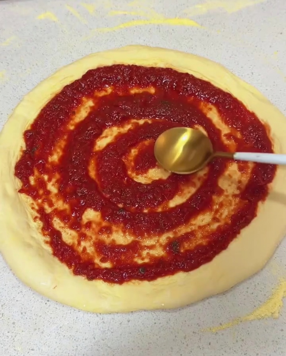 Extendiendo salsa de tomate roja con un movimiento circular sobre una base de masa estirada usando el reverso de una cuchara.