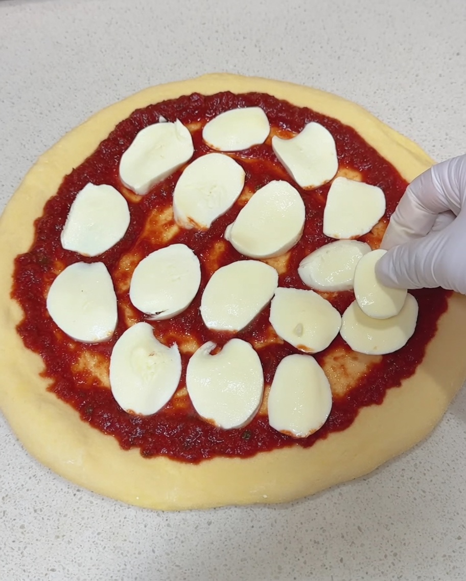 Una mano enguantada colocando rodajas de queso mozzarella fresco sobre una base de pizza cubierta de salsa.