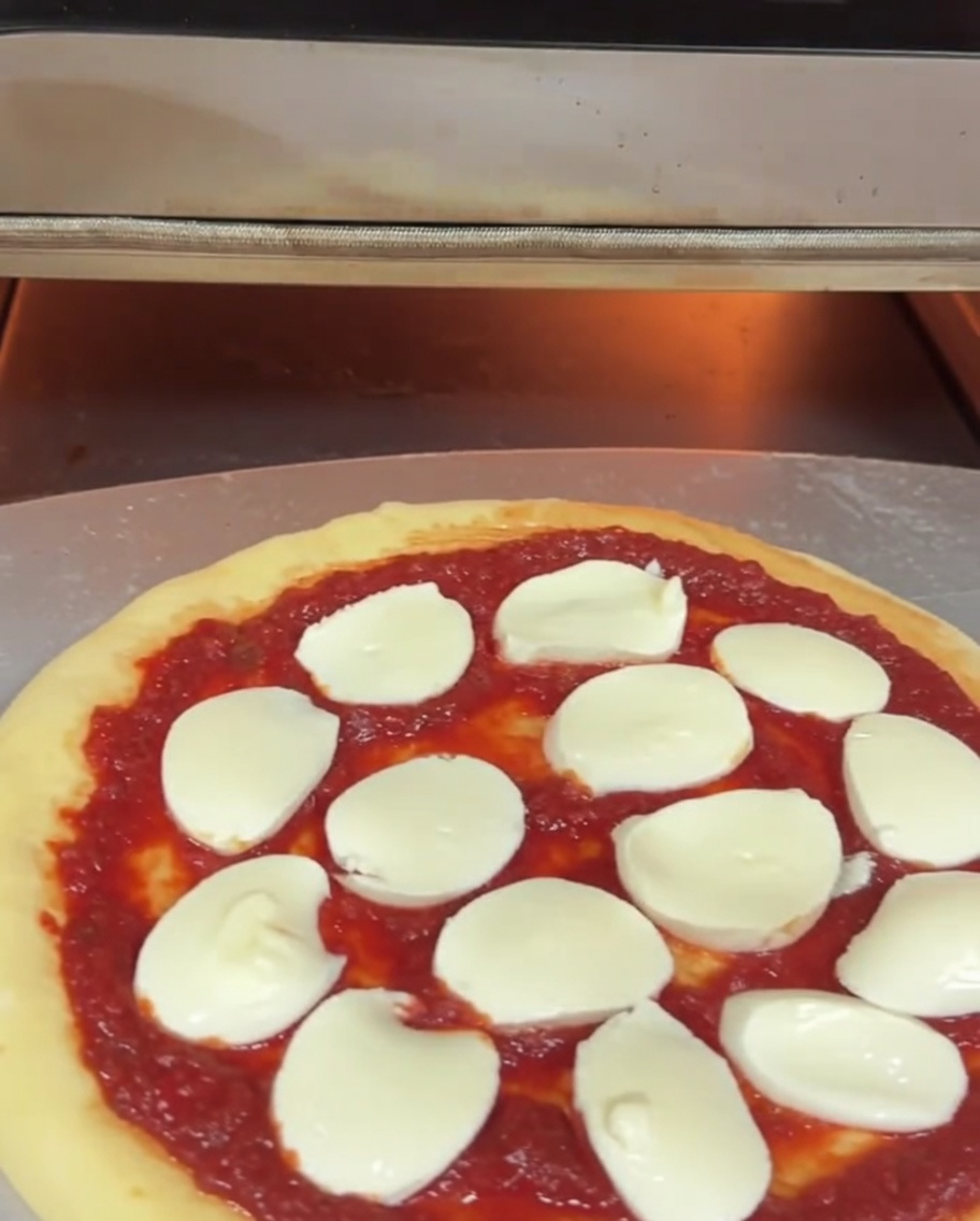 Una pizza cruda con salsa y mozzarella siendo introducida en un horno eléctrico para pizza con una pala metálica.