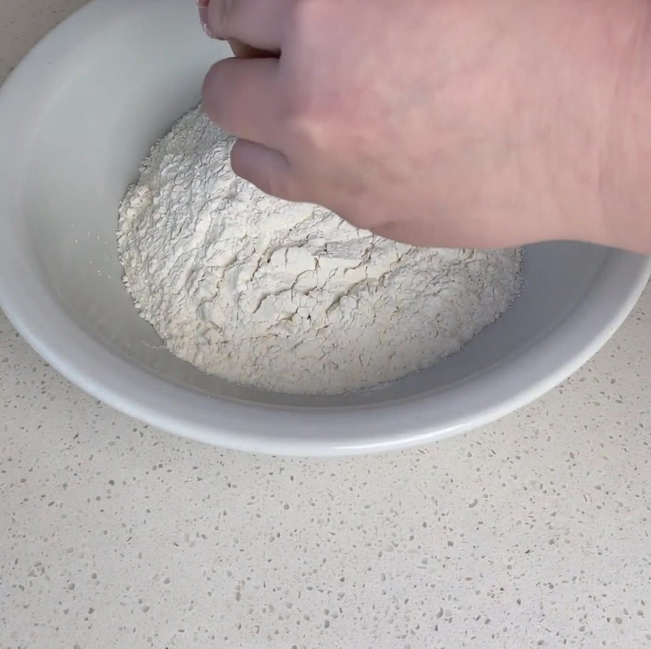 Manos rompiendo un huevo crudo en un volcán de harina en un bol.