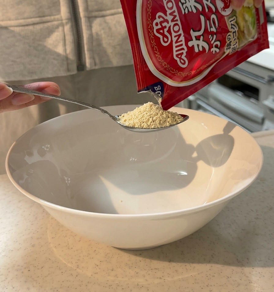 Polvo de caldo de pollo siendo vertido desde una cuchara en un tazón grande blanco.
