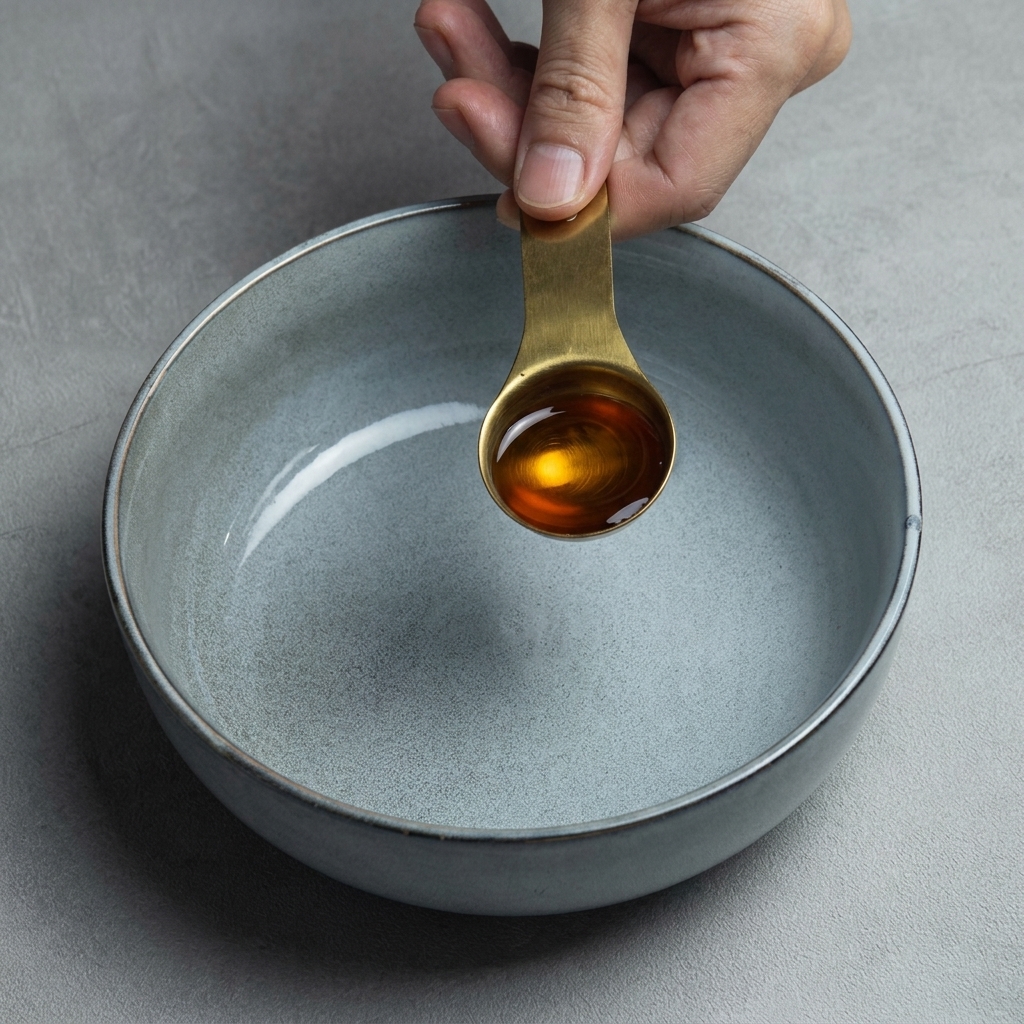 Una mano sosteniendo una cuchara medidora de latón llena de aceite de sésamo oscuro sobre un bol de cerámica gris.