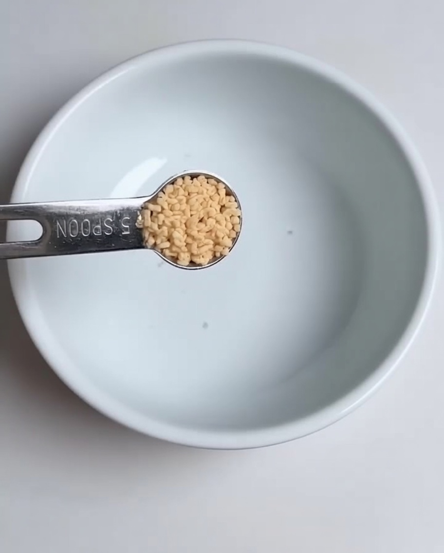 Primer plano de una cuchara medidora añadiendo caldo de pollo en polvo a un cuenco blanco.