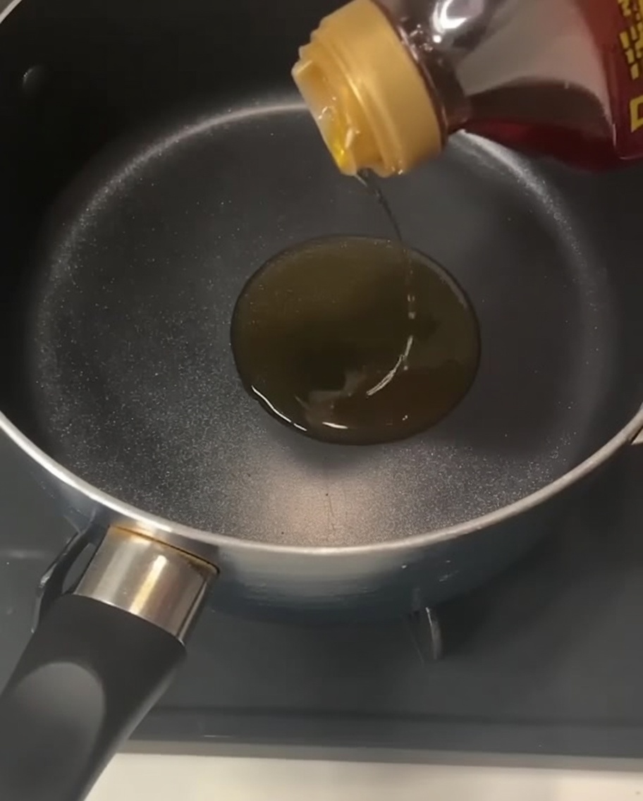 Una botella vertiendo aceite de sésamo dorado y transparente en una olla negra vacía sobre una estufa.