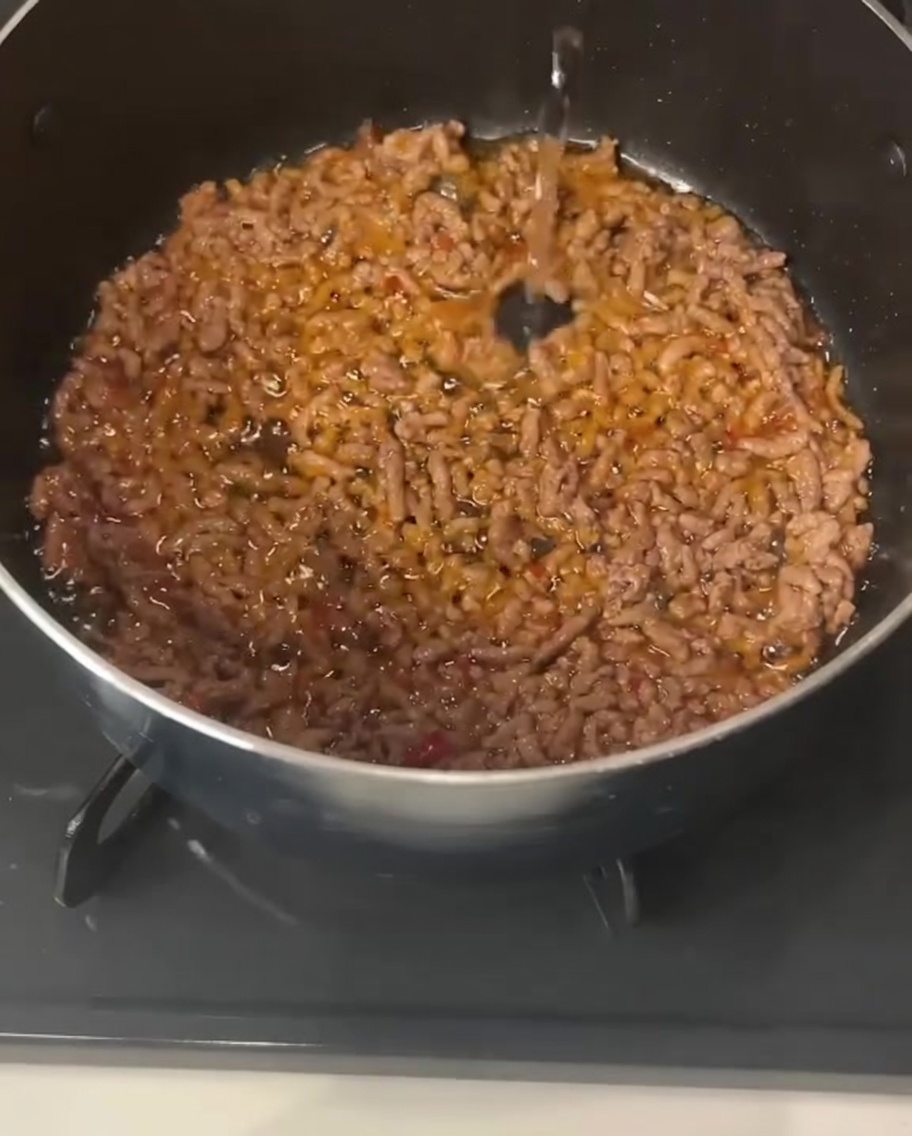 Líquido transparente vertiéndose en una olla que contiene carne picada de cerdo cocinada y sazonada.