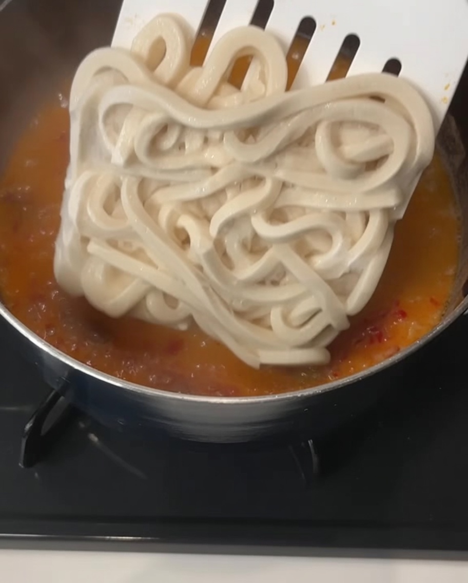 Un bloque grande y cuadrado de fideos udon gruesos congelados siendo introducido por una espátula en un caldo naranja cremoso y burbujeante.