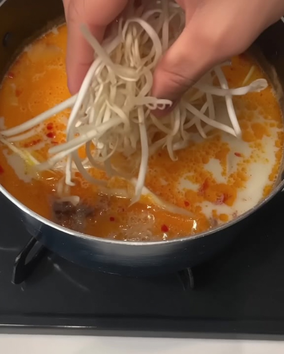 Una mano arrojando un puñado de brotes de soja blancos y frescos en una olla de sopa de fideos cremosa.