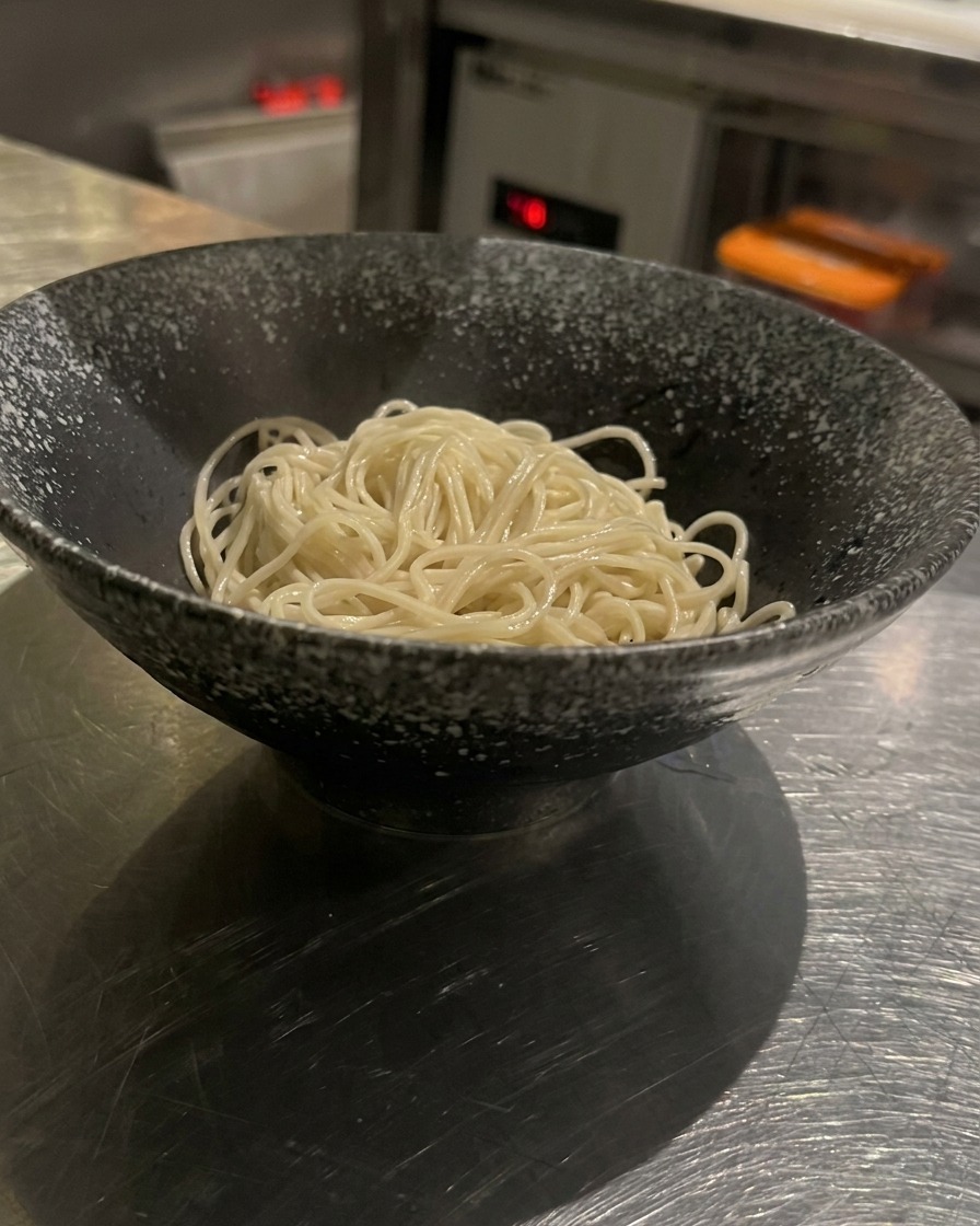 Fideos cocidos en un tazón negro siendo cubiertos con caldo caliente rico y cremoso.