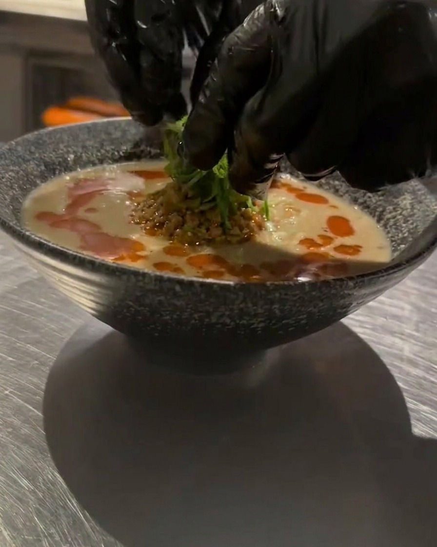 Manos con guantes colocando cebolleta picada sobre un tazón de Tan Tan Ramen caliente.