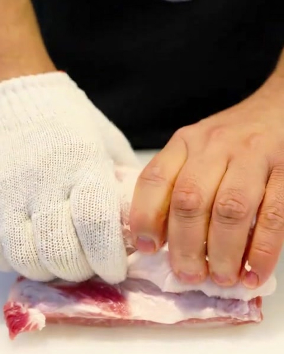 Un rollo de panceta de cerdo siendo atado firmemente con hilo de cocina por manos con guantes protectores.