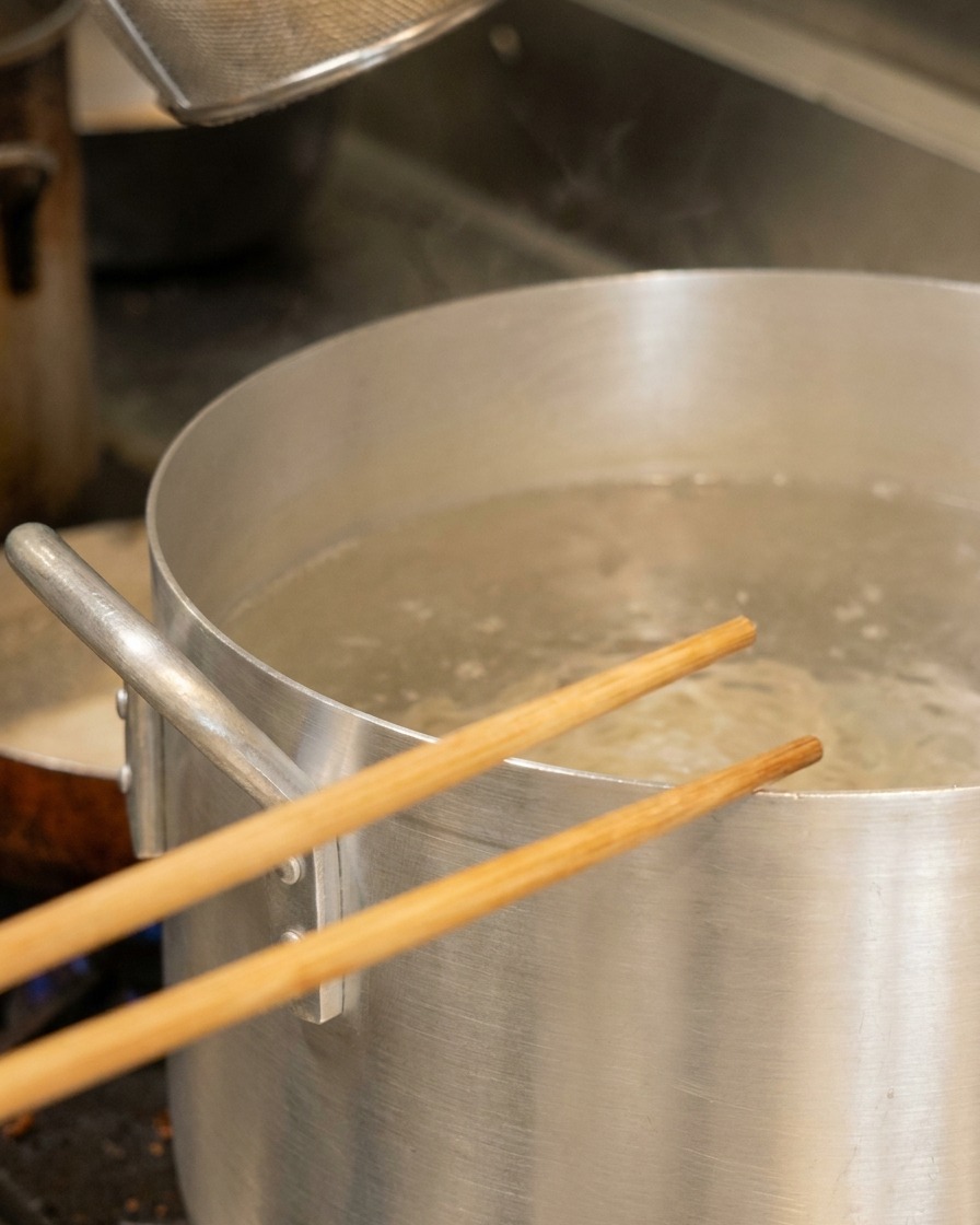 Palillos de madera descansando sobre una olla grande de aluminio con agua hirviendo y una cesta para fideos.