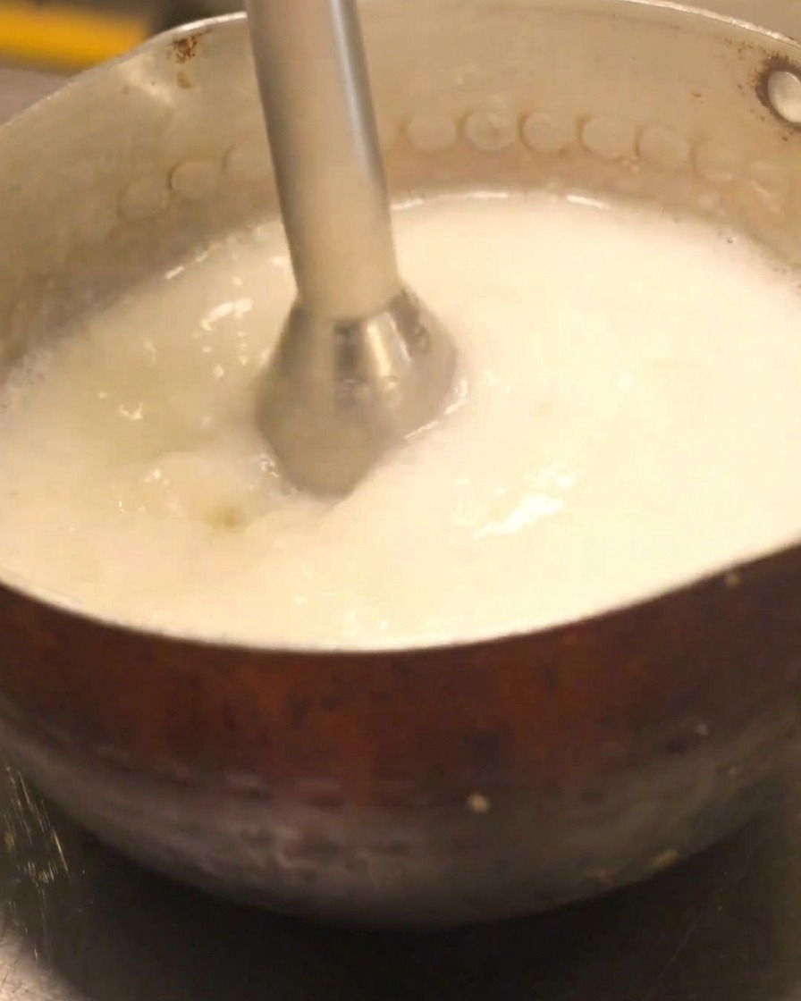Una batidora de mano siendo usada para crear una espuma cremosa en una olla de caldo de pollo paitan.