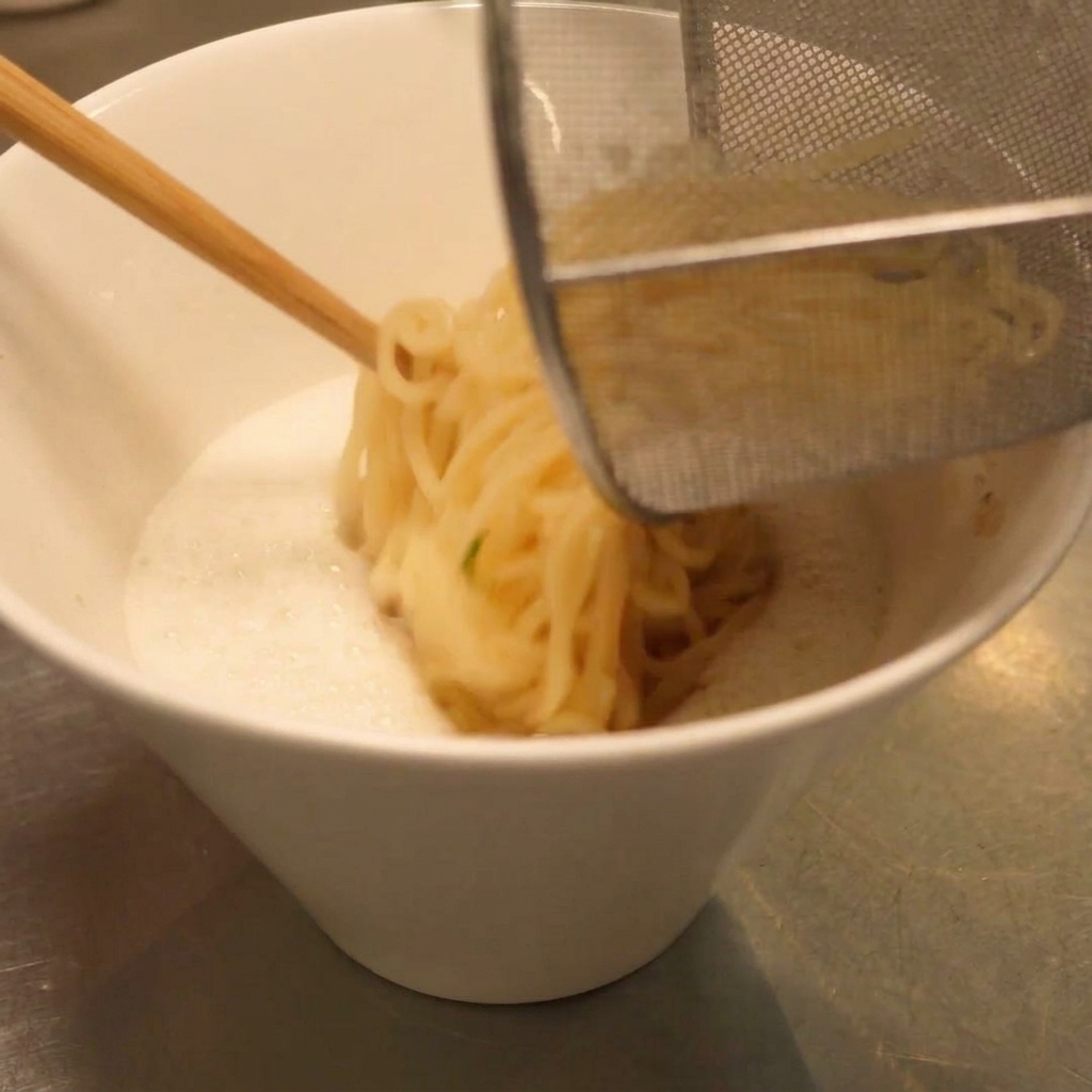Fideos de ramen escurridos siendo transferidos al tazón de sopa cremosa usando una cesta de cocina.