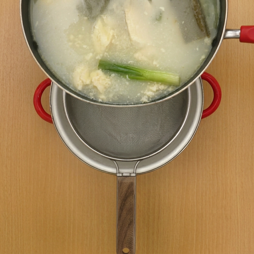 Caldo de pollo caliente siendo vertido a través de un tamiz metálico hacia una olla limpia.
