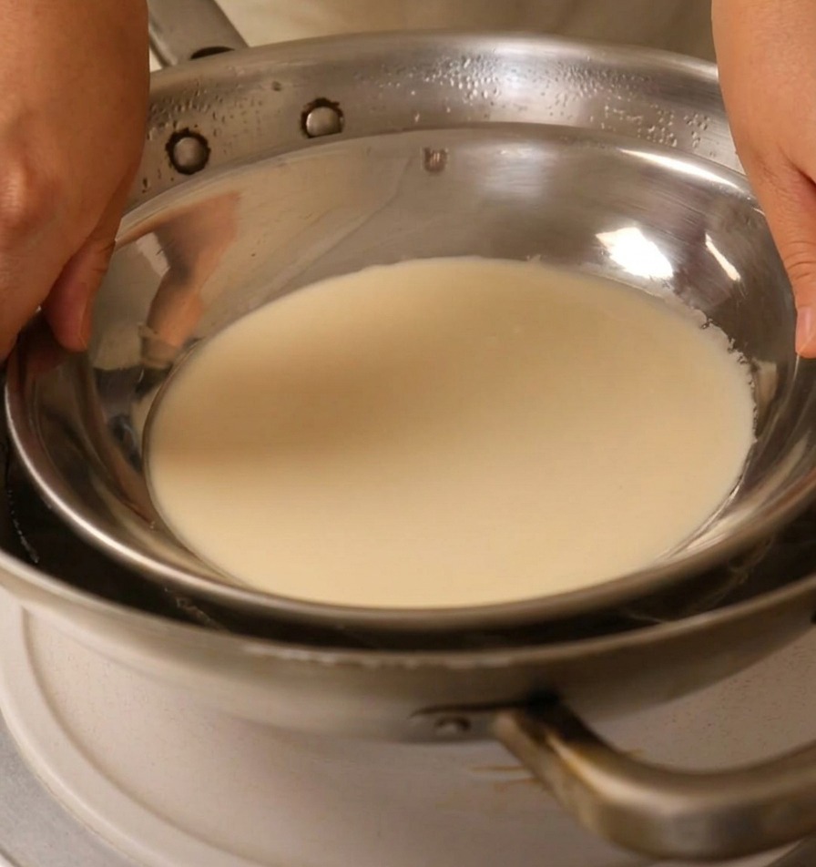 Manos colocando un plato de metal lleno de masa blanca sobre un wok con agua hirviendo.