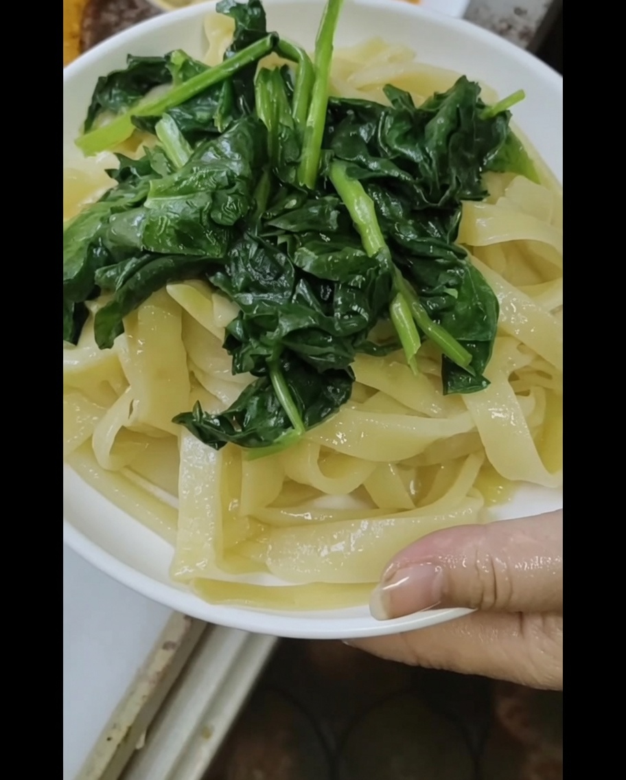 Vista en primer plano de fideos recién hechos al vapor cubiertos con espinacas blanqueadas, siendo rociados con una rica salsa de sésamo y cacahuete.