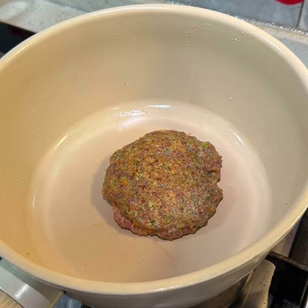 Una hamburguesa de carne sazonada cocinándose en una sartén, mostrando una superficie dorada.