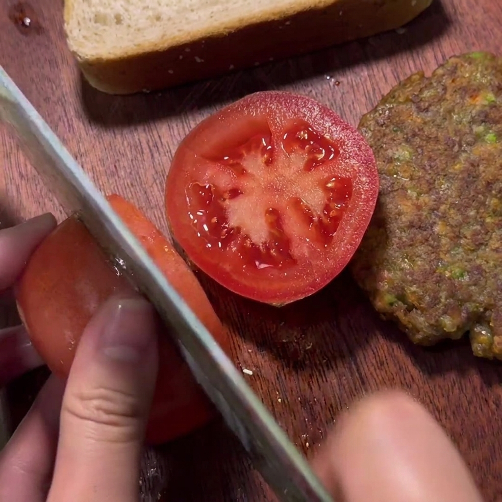 Una mano cortando un tomate rojo fresco en una tabla de madera junto a una hamburguesa de carne cocida.