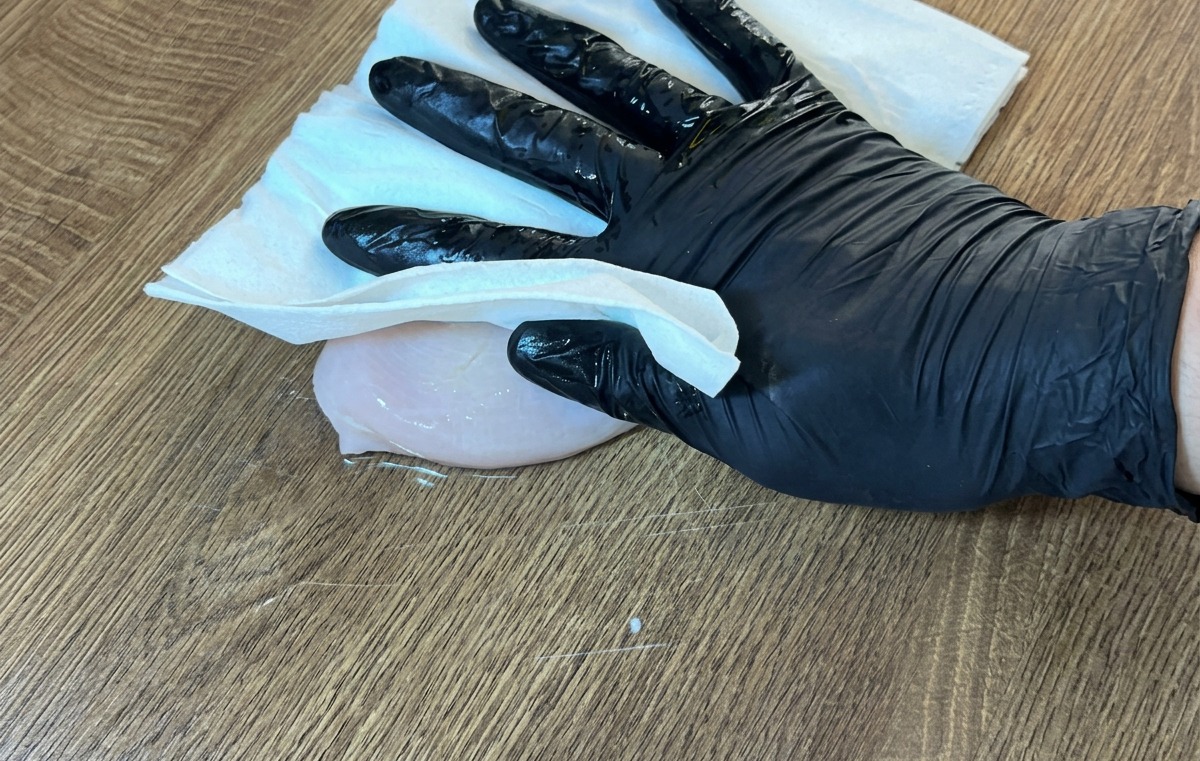 Mano enguantada usando papel de cocina para secar una pechuga de pollo cruda sobre una superficie de madera.