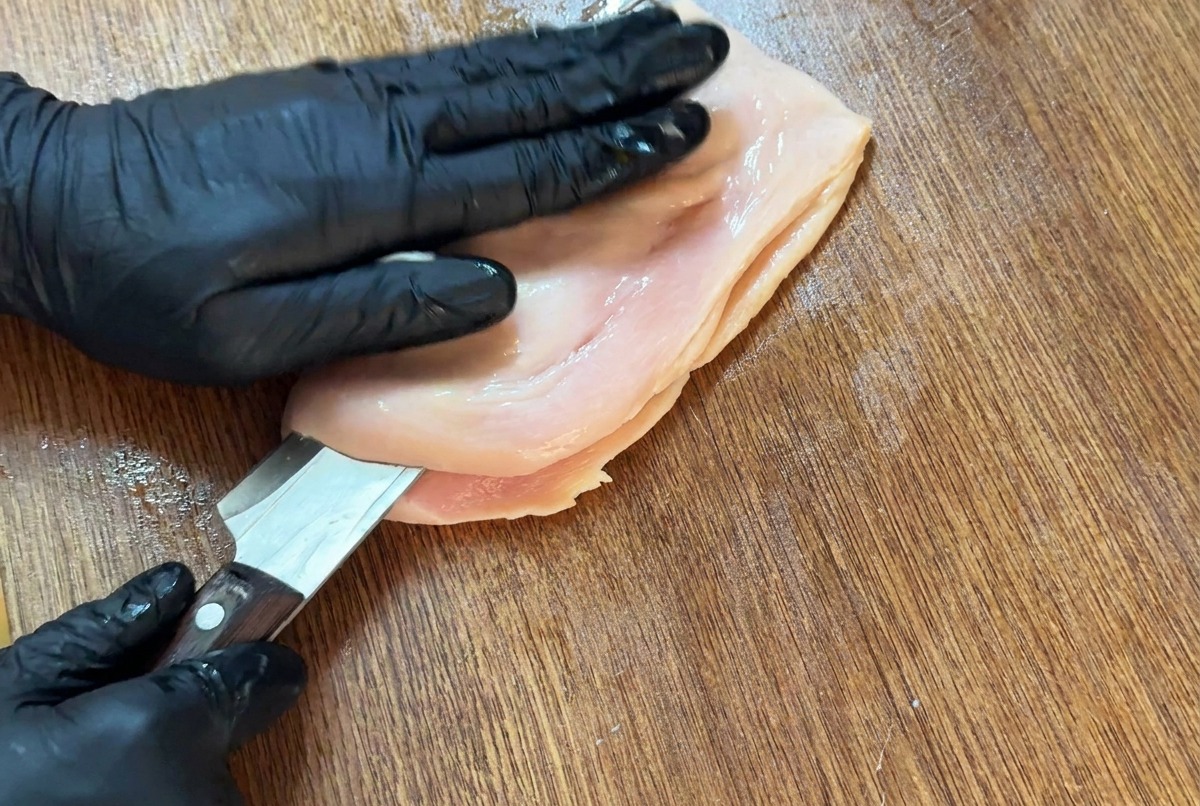 Cuchillo afilado cortando horizontalmente una pechuga de pollo sobre una tabla para abrirla en forma de mariposa.