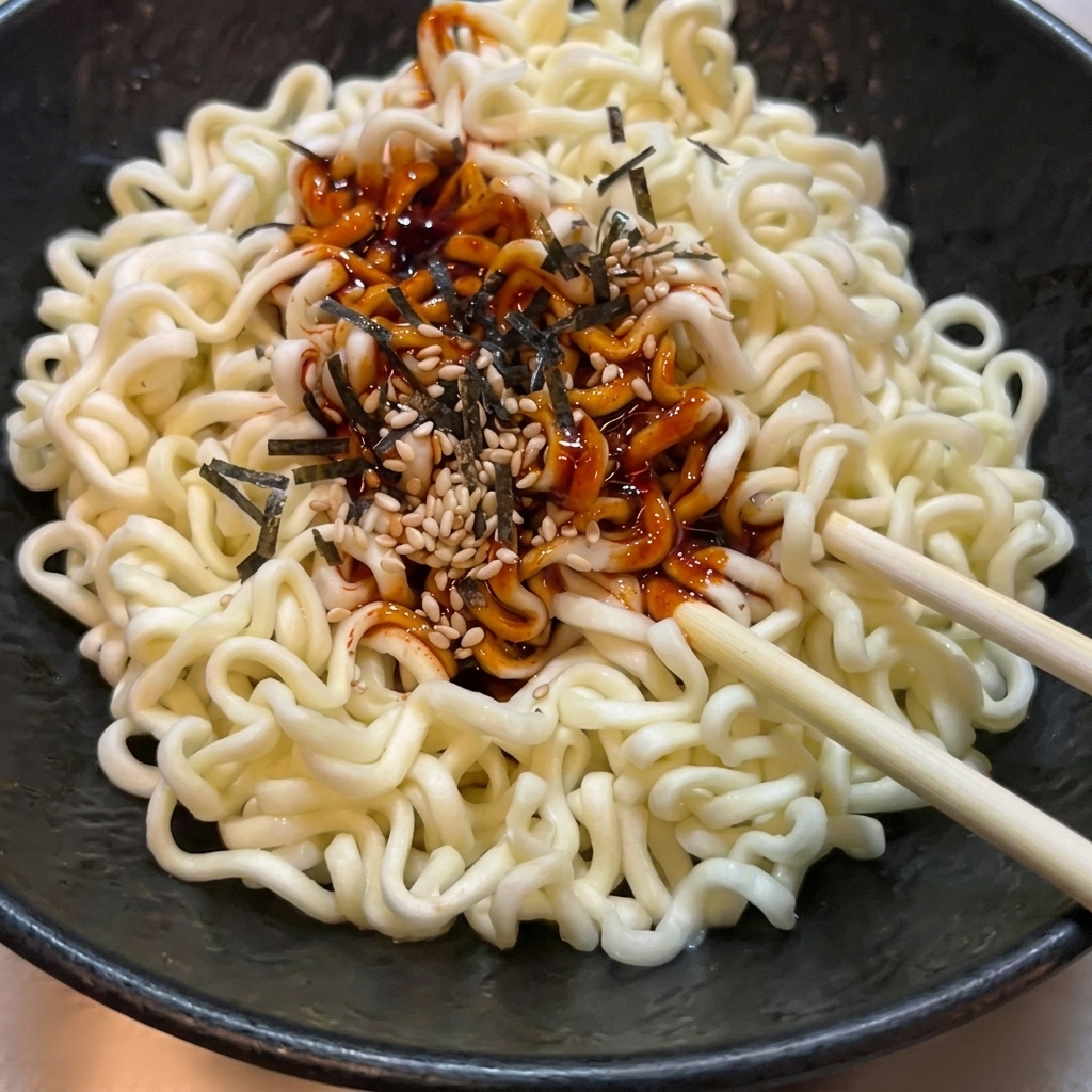 Fideos ramen ondulados en un bol negro siendo mezclados con salsa picante roja, semillas de sésamo y algas con palillos de madera.